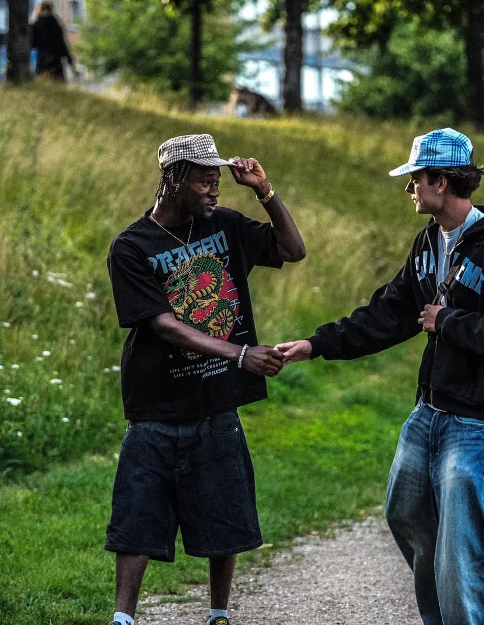 Two guys shaking hands in a park, one in the Tobacco cap